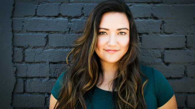 A captivating portrait of a woman smiling confidently in front of a brick wall, showcasing her long brunette hair and vibrant eyes.