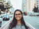 A young woman with glasses smiling on a city street, embracing urban lifestyle.